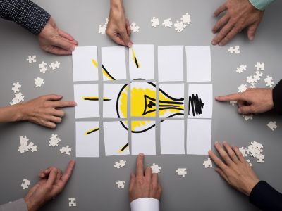 Top view of businesspeople hands touching white papers arranged on a gray table forming a yellow light bulb. Conceptual for brainstorming and teamwork.